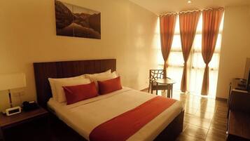 Quarto de hotel com uma cama grande, mesa de cabeceira com luminária, uma cadeira e uma escrivaninha.