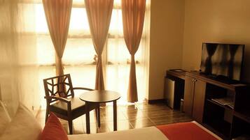 Quarto de hotel com cama, televisão, mesa pequena, cadeira e cortinas.