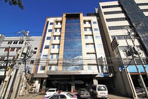 Un edificio de varios pisos con fachada de vidrio central, un letrero que dice "VERJANDEL" y varios vehículos estacionados al frente.