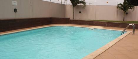 A rectangular swimming pool with a tiled edge, surrounded by a low wall and a grassy area with palm trees.