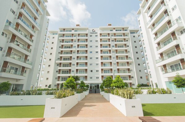 Un edificio residencial de varios pisos con balcones, una entrada central y áreas ajardinadas con árboles.