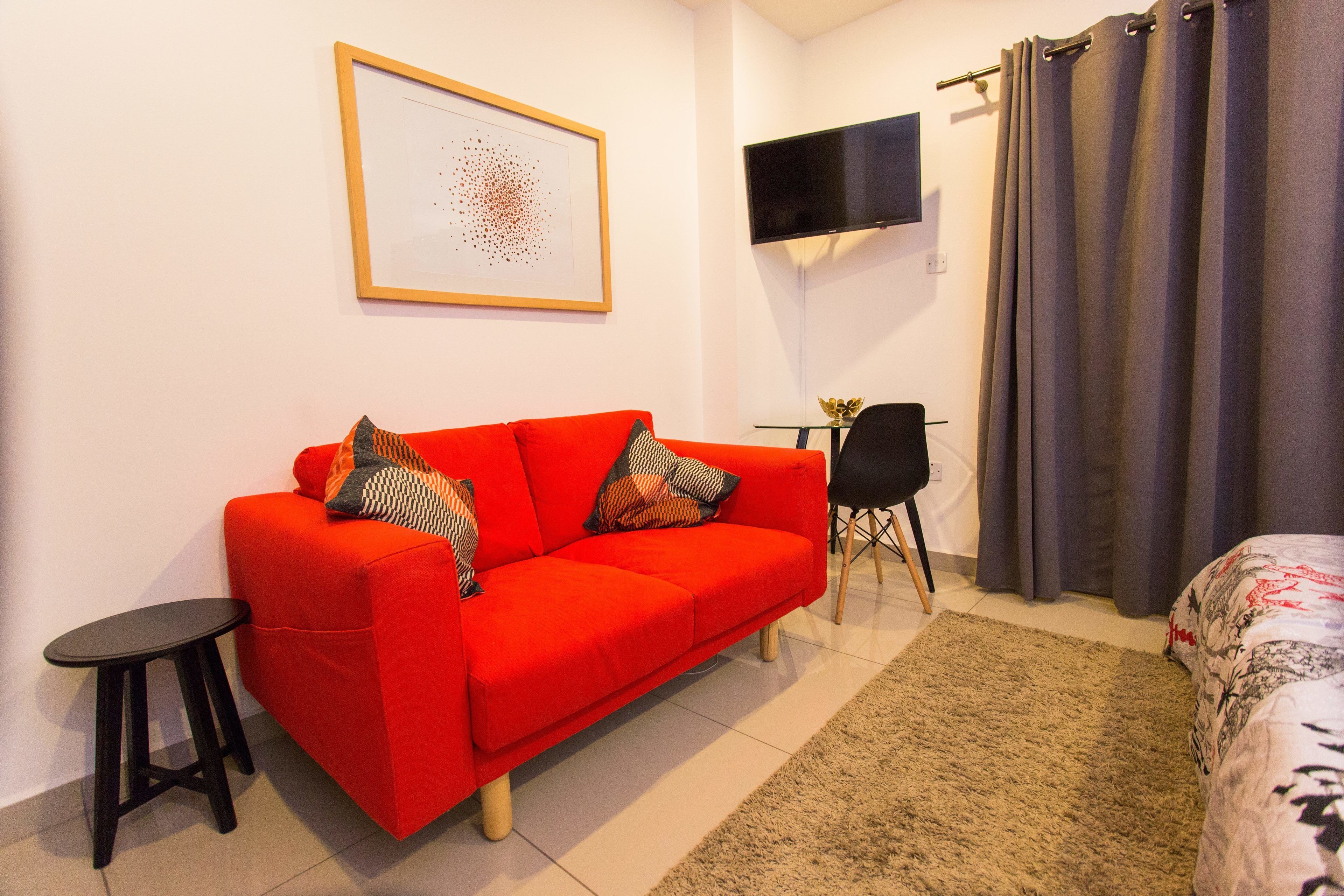 Een moderne woonkamer met een rode bank, een klein zwart tafeltje, een aan de muur bevestigde televisie en een grijze gordijn.