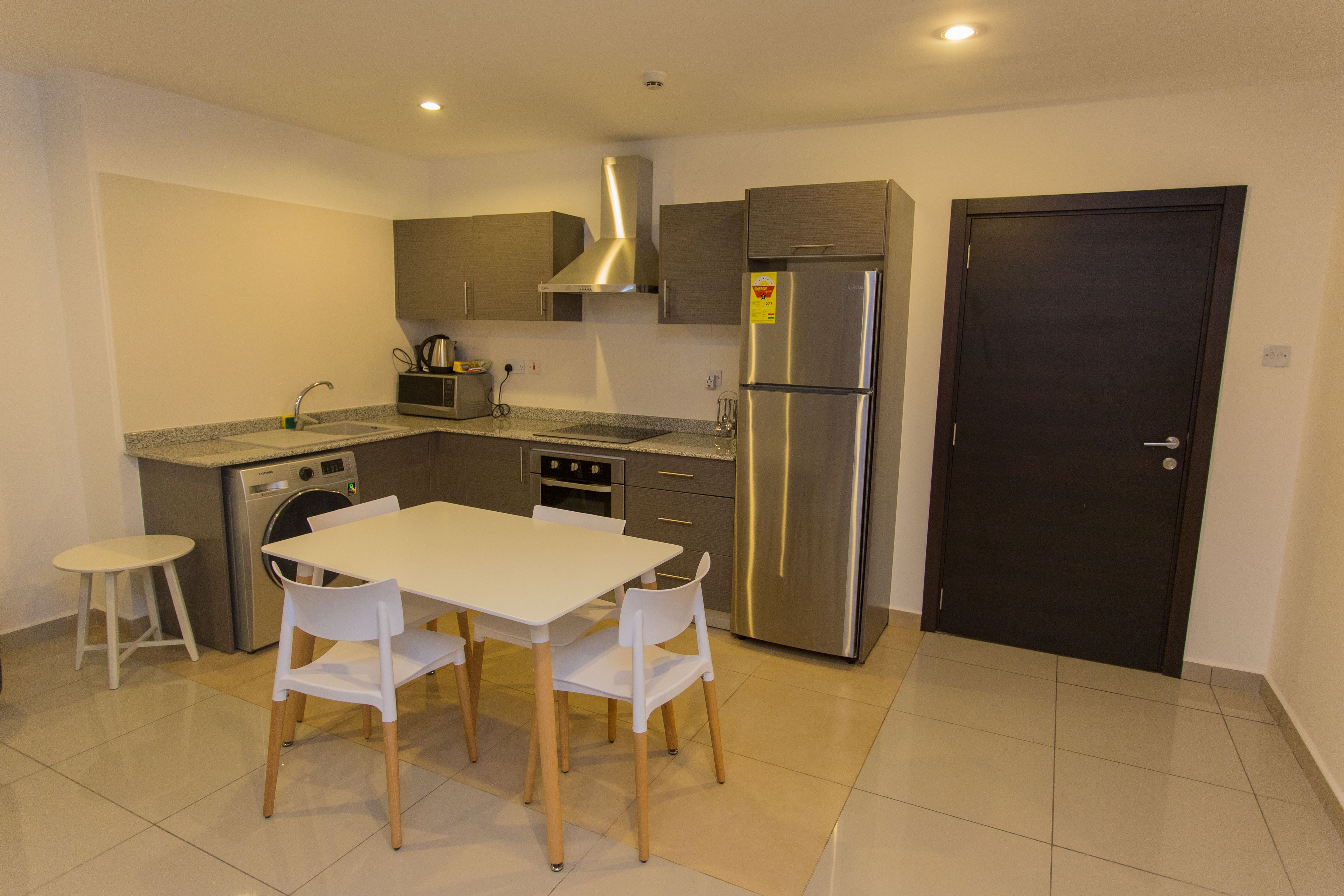 A compact kitchen with a stainless steel refrigerator, a built-in oven, and a washing machine. There is a dining table with four chairs and a small stool.