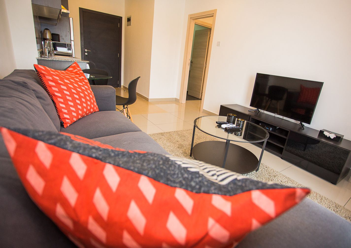 Una sala moderna con un sofá gris, cojines rojos estampados, una mesa de centro de vidrio, un televisor de pantalla plana y una zona de cocina al fondo.