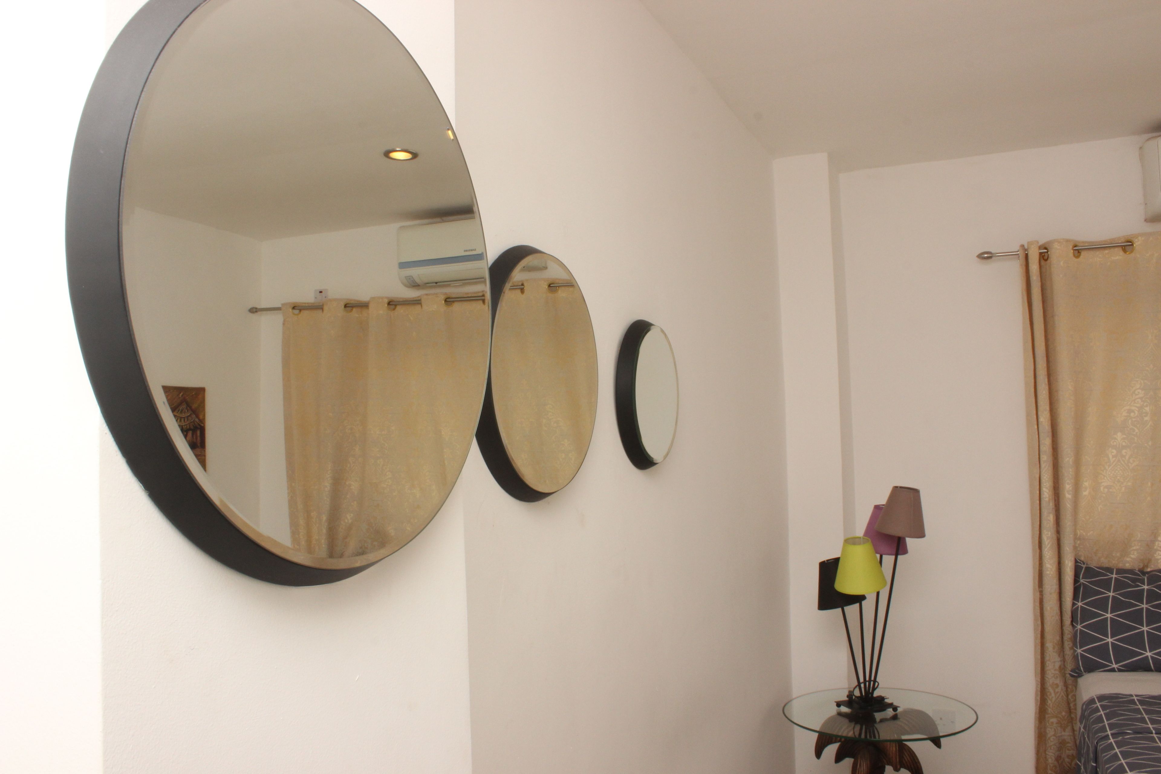 A room with three round mirrors on the wall, a glass table with a lamp, and a bed with a patterned bedspread.
