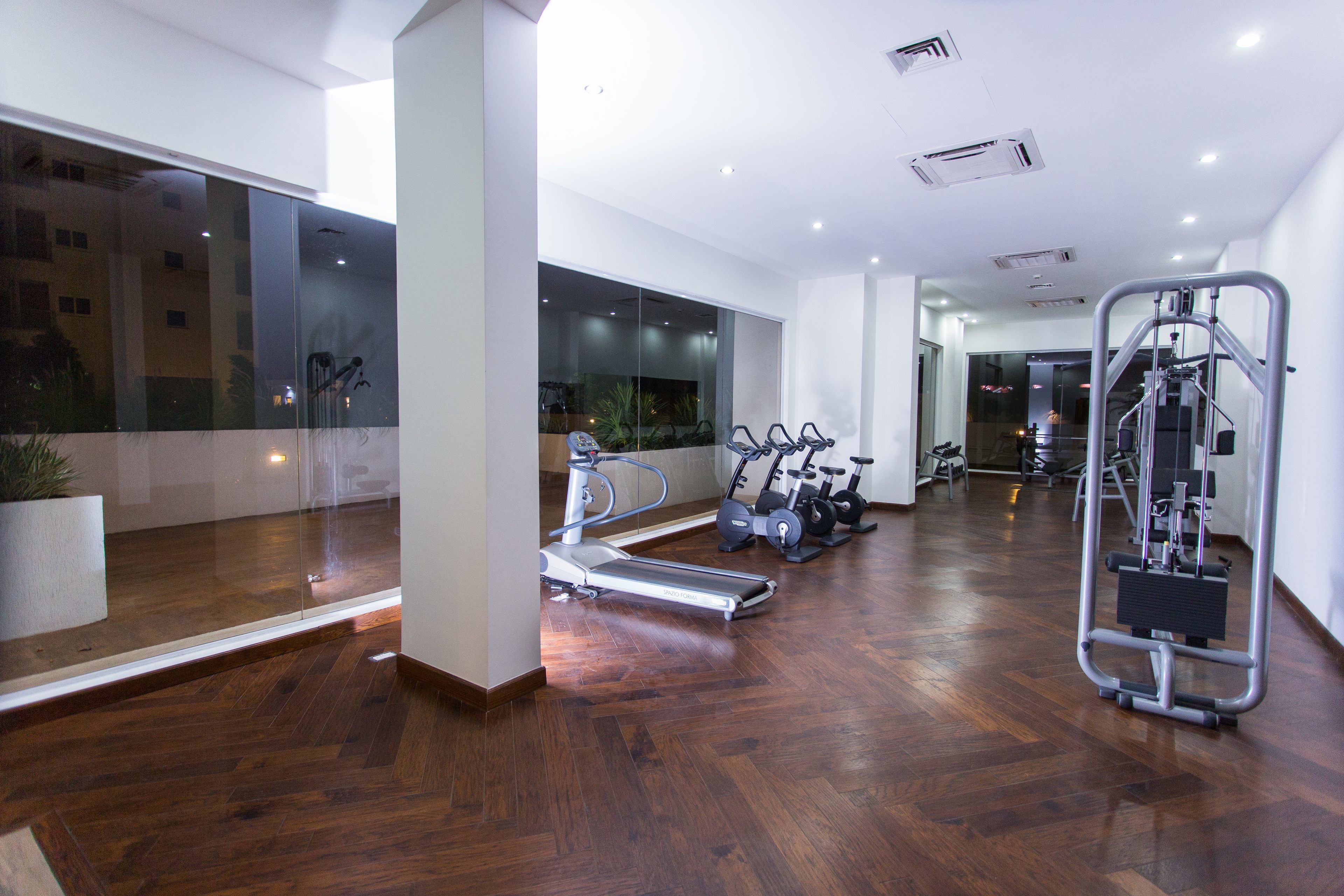A modern gym with exercise equipment, large windows, and a wooden floor.