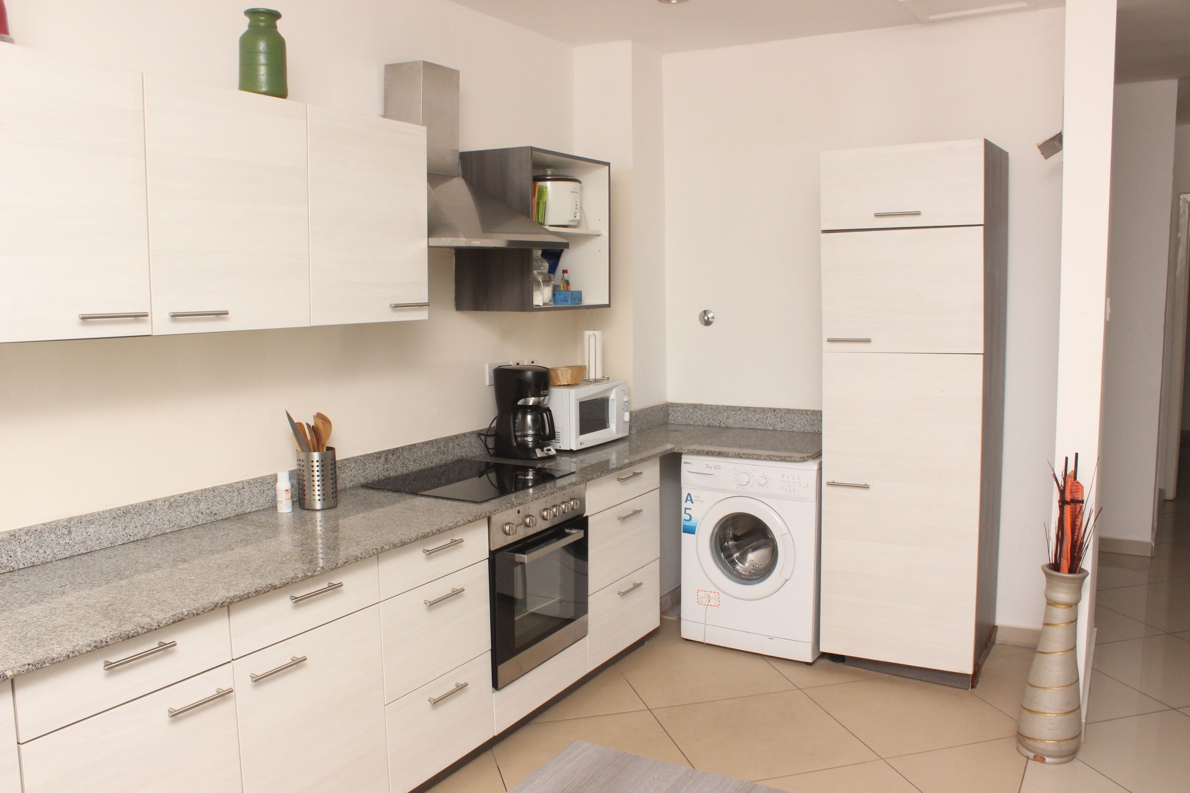 Cocina compacta con armarios blancos, microondas, cafetera y lavadora.