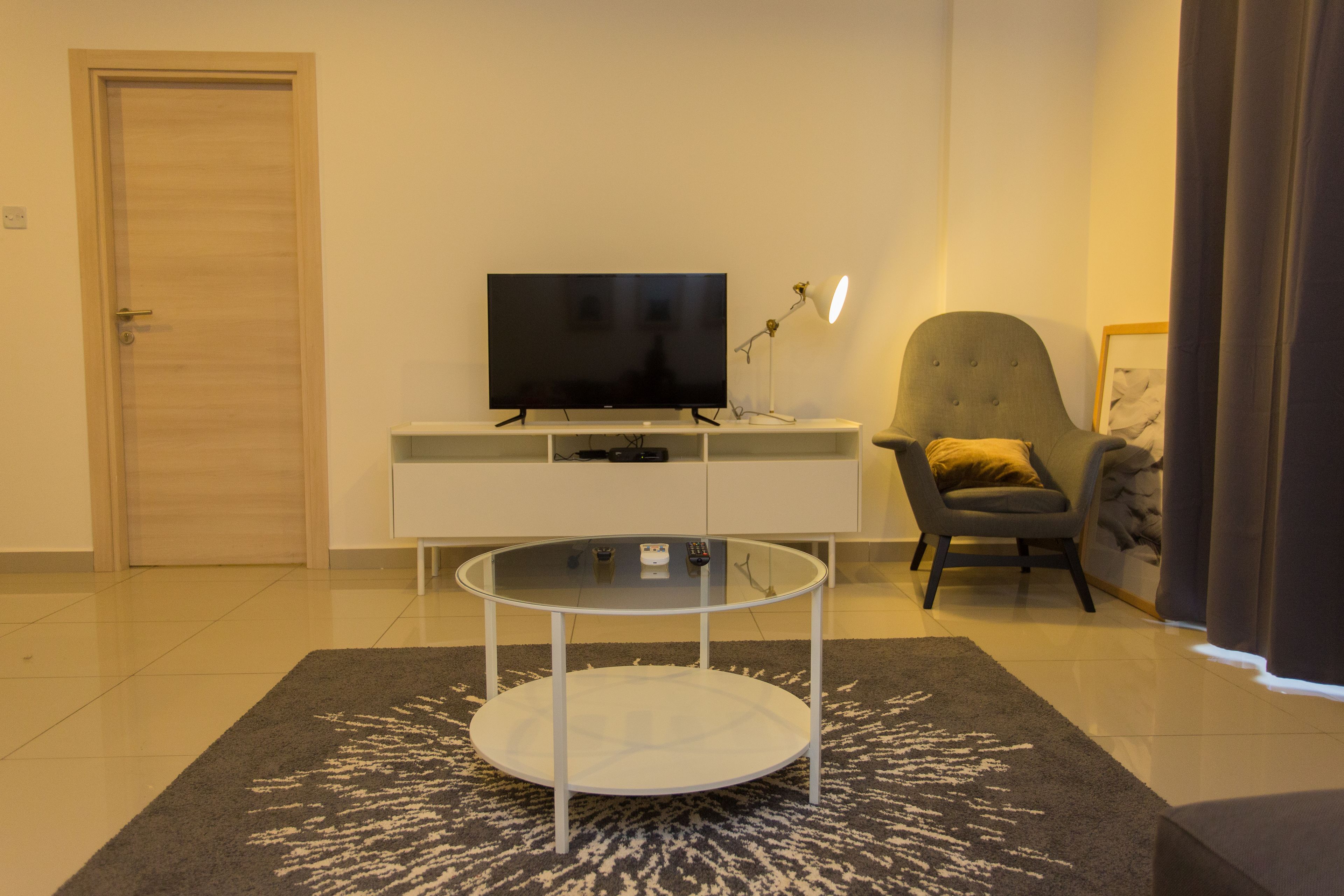 A modern living room with a flat-screen TV on a white console table, a glass coffee table, a grey armchair, and a dark patterned rug.