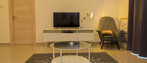 A modern living room with a flat-screen TV on a white console table, a glass coffee table, a grey armchair, and a dark patterned rug.