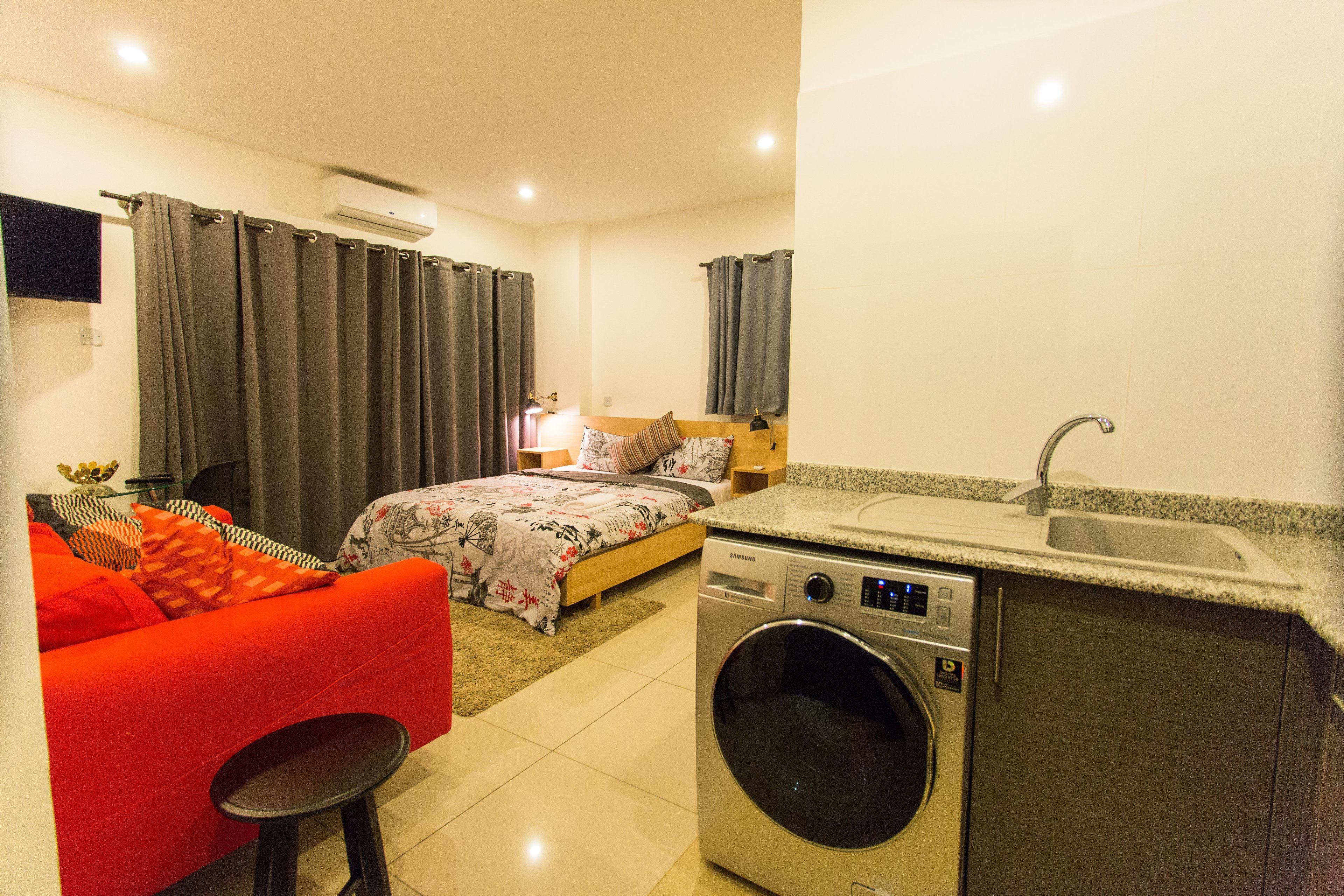 A compact hotel room with a bed, a red sofa, a washing machine, and a sink.