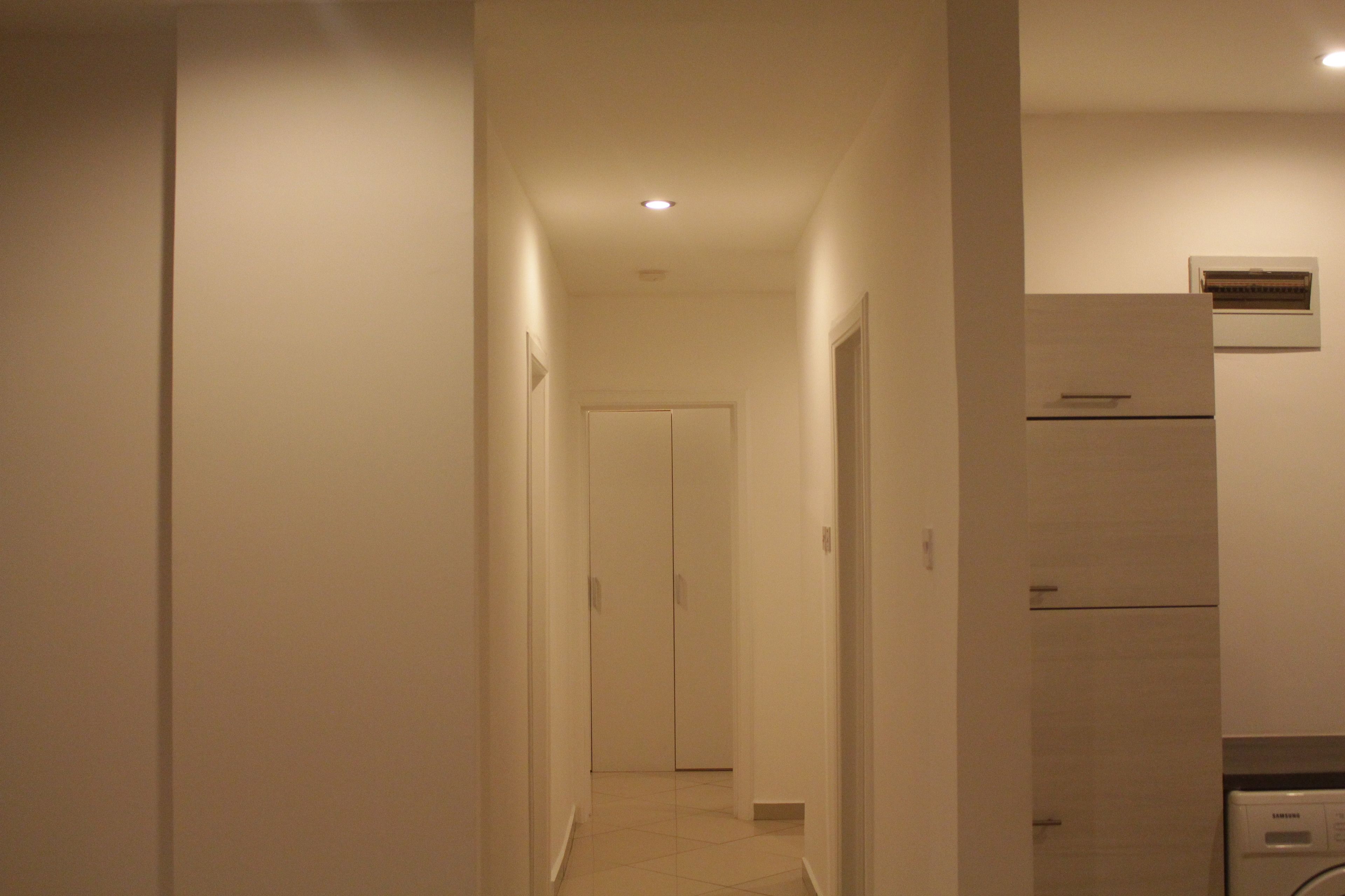 Un pasillo estrecho con una puerta al final, un aire acondicionado de pared y una zona de cocina con refrigerador.