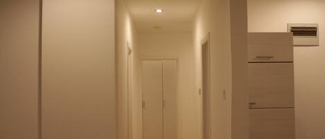 Un pasillo estrecho con una puerta al final, un aire acondicionado de pared y una zona de cocina con refrigerador.