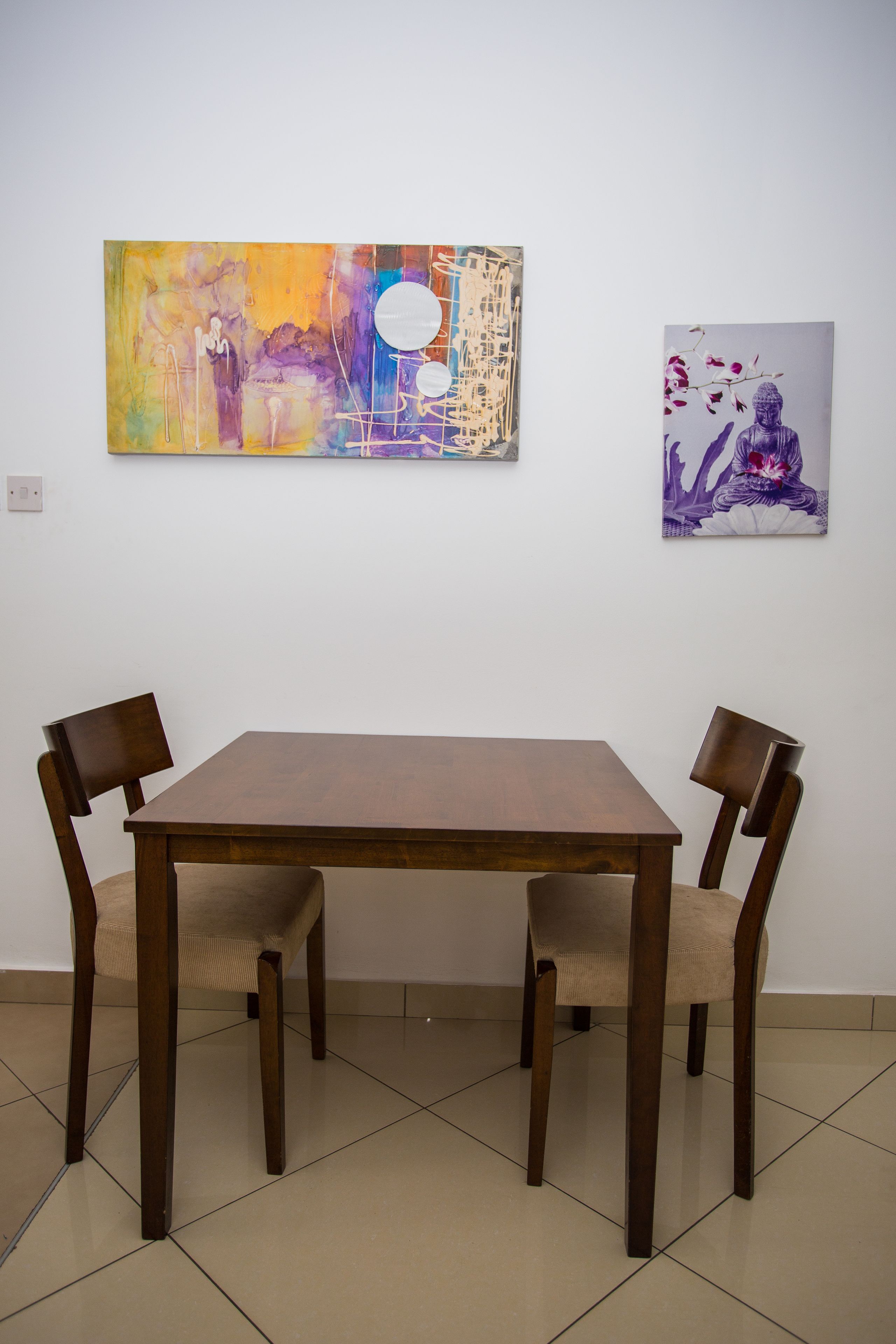A dining area with a wooden table and chairs, two artworks on the wall, and a tiled floor.