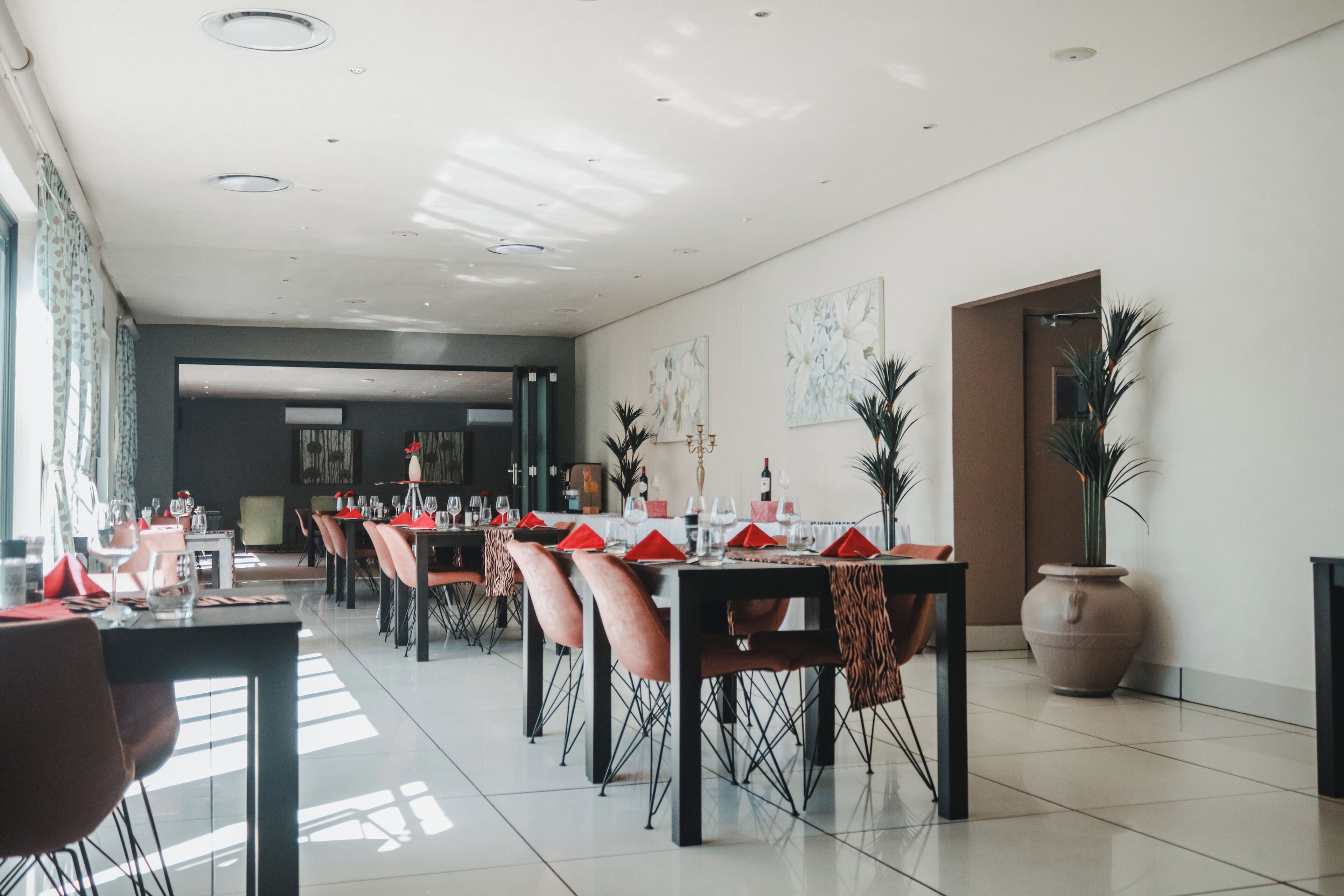 Un moderno comedor con una larga mesa dispuesta para una comida, rodeada de sillas, una planta grande en maceta y obras de arte en las paredes.
