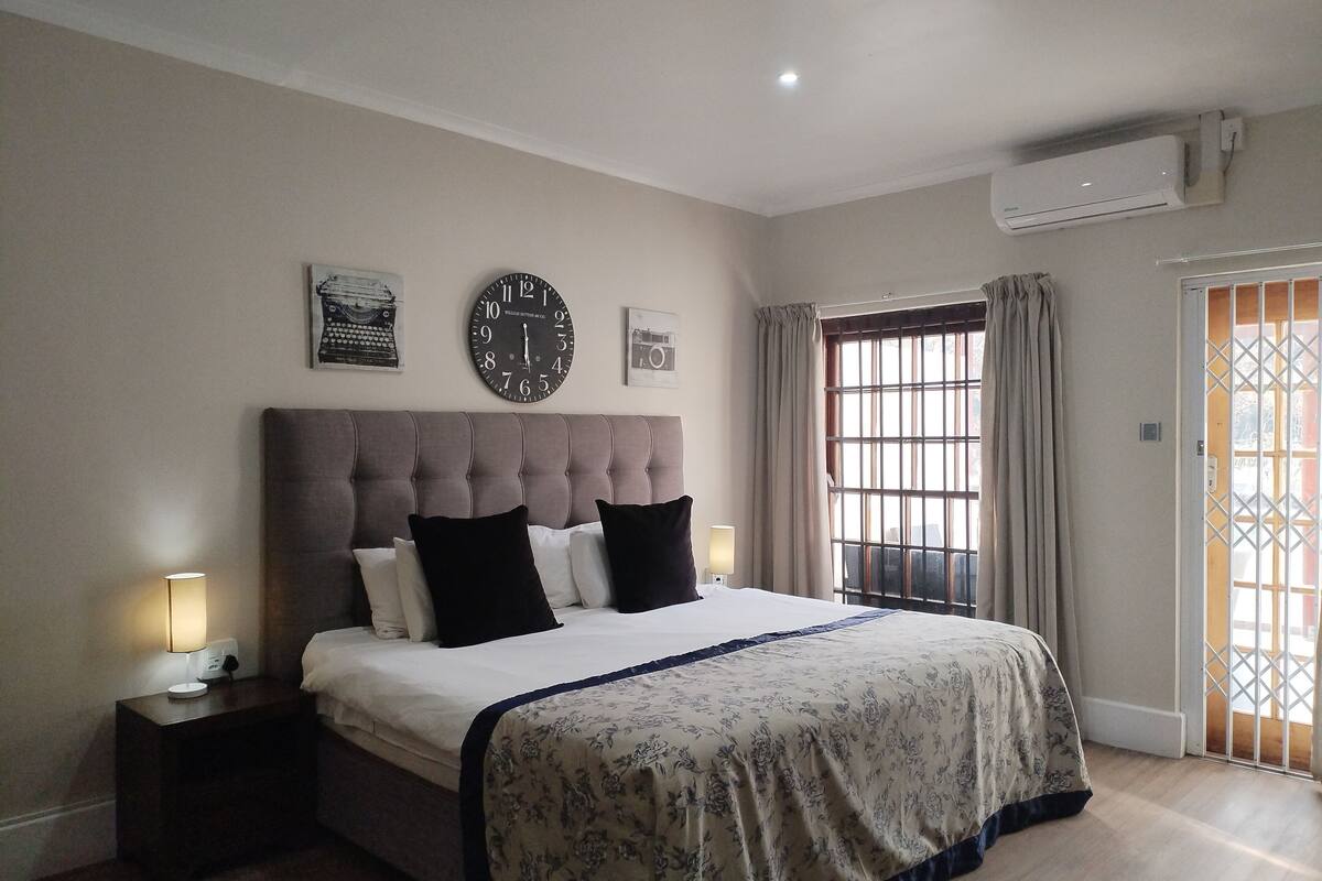 Ein Schlafzimmer mit einer gepolsterten Kopfteile, einem Bett mit einer blumigen Tagesdecke, zwei schwarzen Kissen, einer Wanduhr und einem Fenster mit Vorhängen.