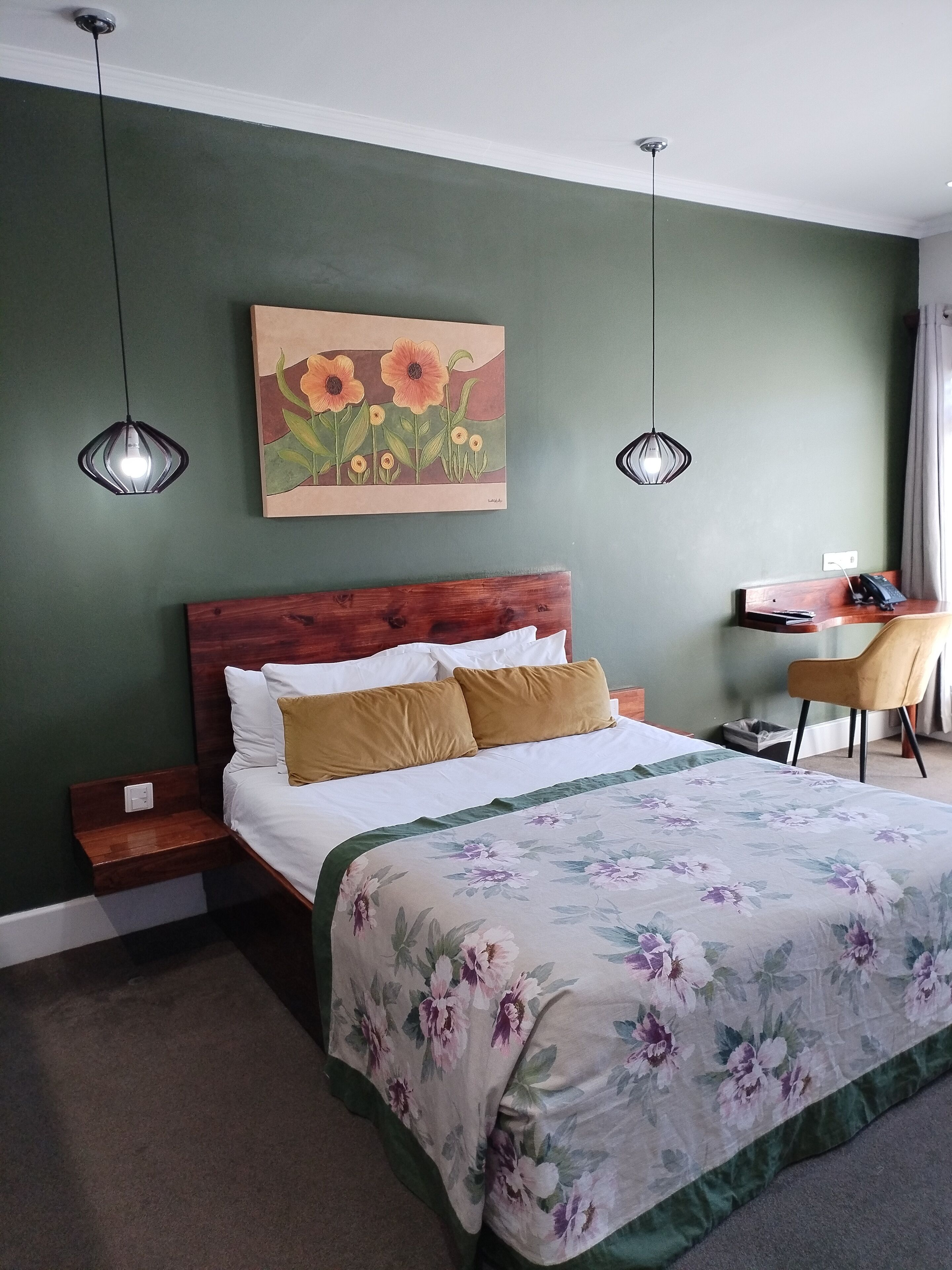 A neatly made bed with a floral bedspread, a wooden headboard, and two bedside tables with lamps. A painting of flowers hangs above the bed, and a desk with a chair is visible in the corner.