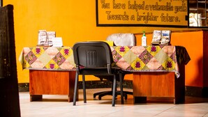 A room with a wooden desk, a black chair, and a table covered with a colorful quilt. A sign on the wall reads, "And leave a trace of your passage. You have helped a vulnerable youth towards a brighter future."