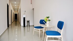 Un couloir avec une table blanche et deux chaises bleues, une poubelle et un meuble blanc.