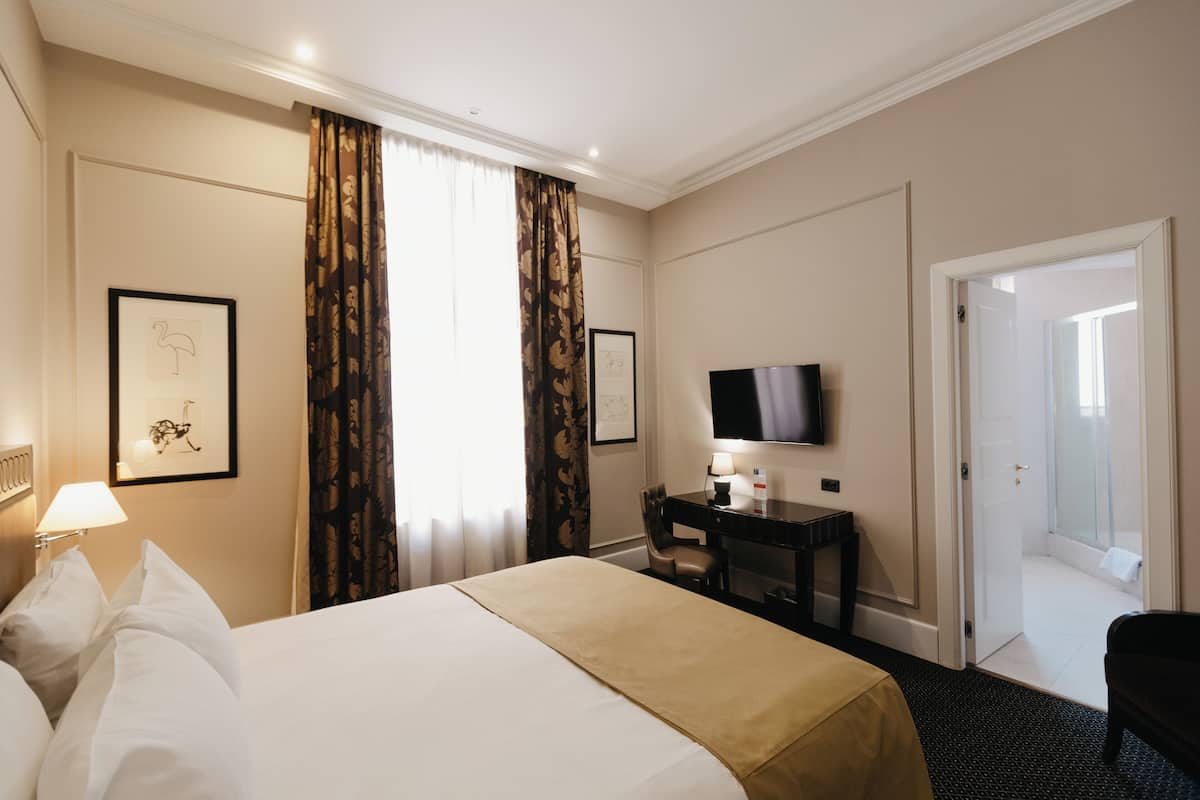 A hotel room with a bed, a desk, a chair, a TV, and a door leading to a bathroom.