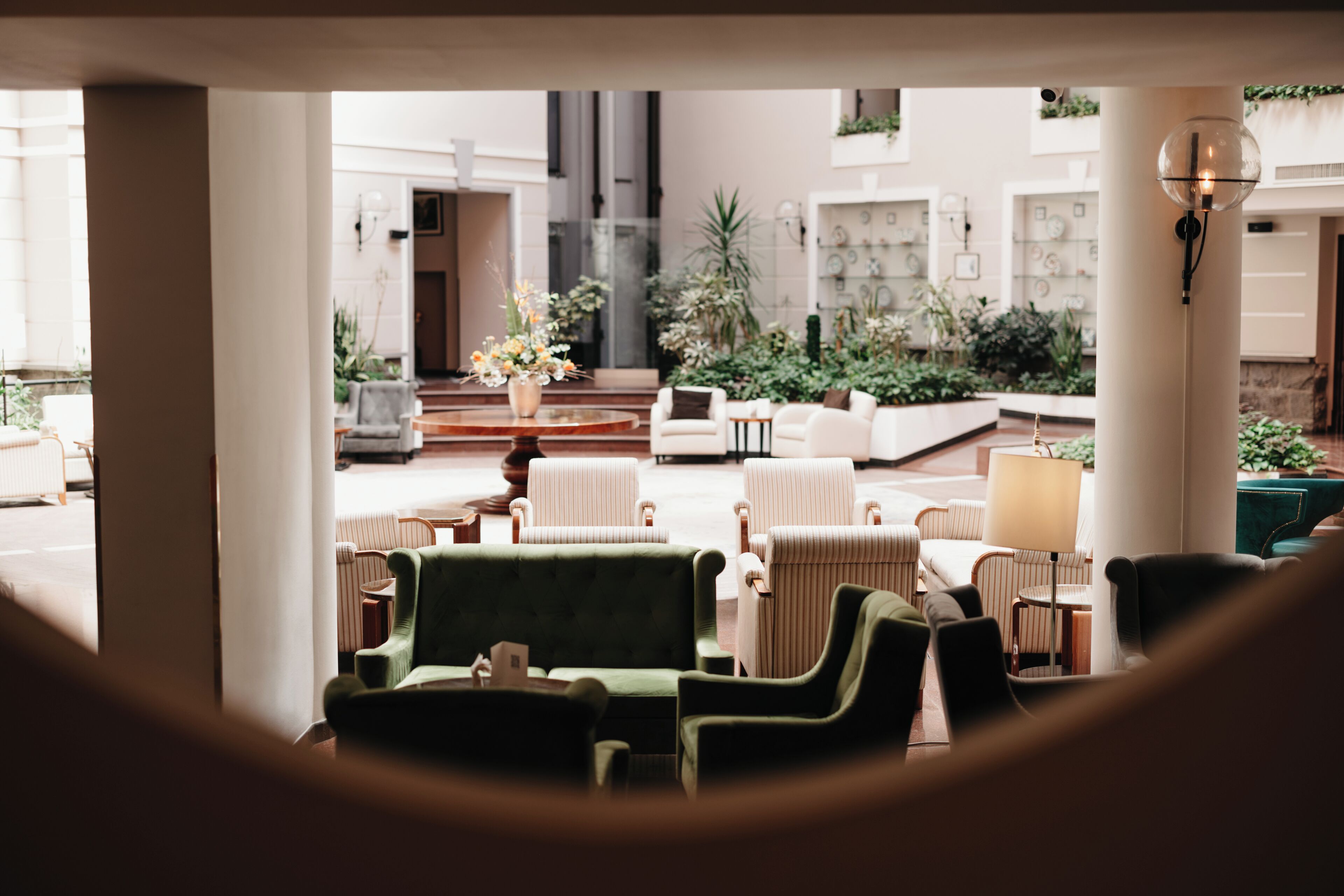 Een moderne lobby met een groene bank, witte fauteuils en een salontafel. Er staan potplanten en een grote vaas met bloemen.