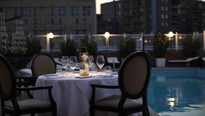 A dining table set for two with chairs, a pool in the background, and buildings in the distance. - Grand Hotel Yerevan (Yerevan)
