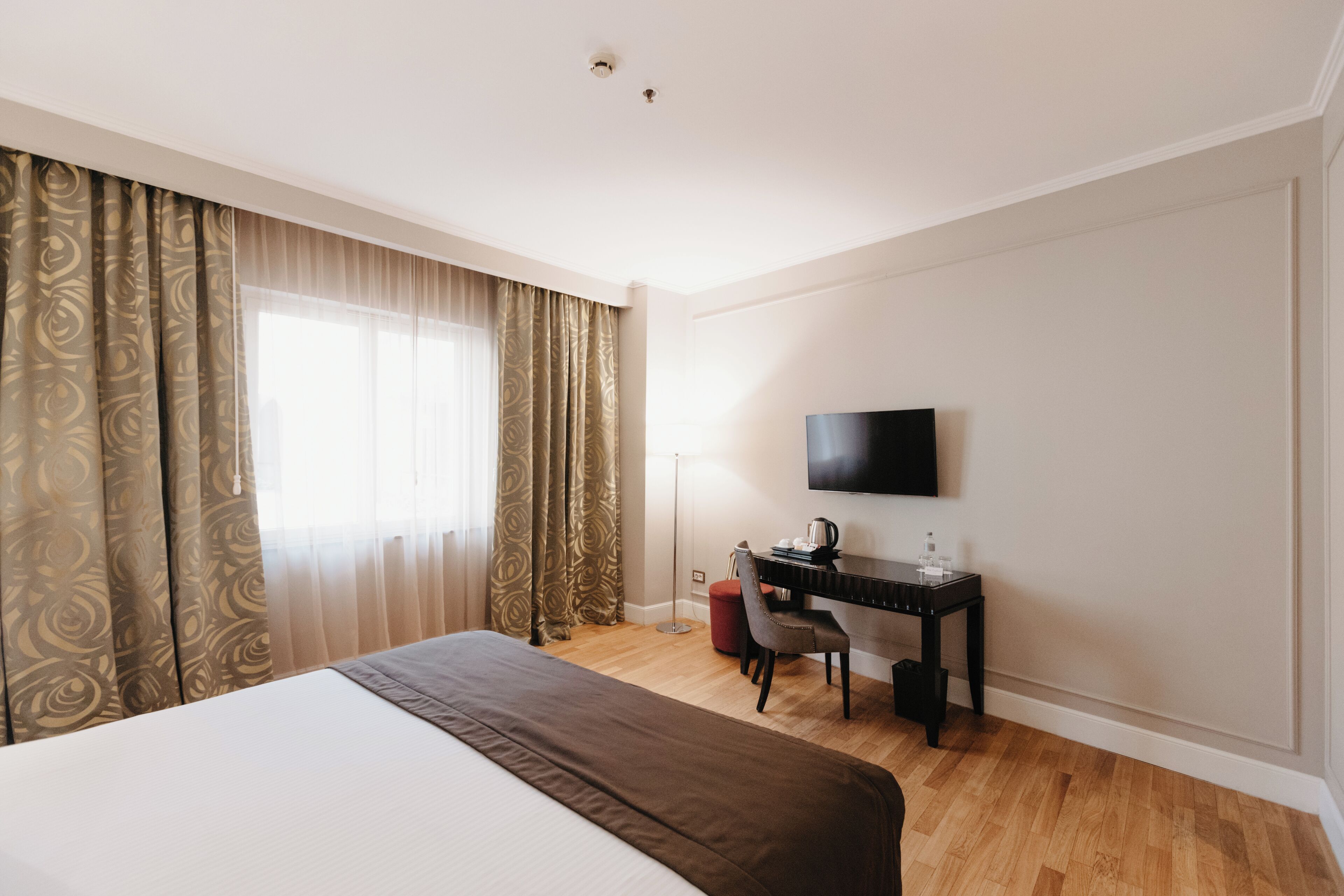A hotel room with a large bed, a desk with a chair, a flat-screen TV, and a window with curtains.