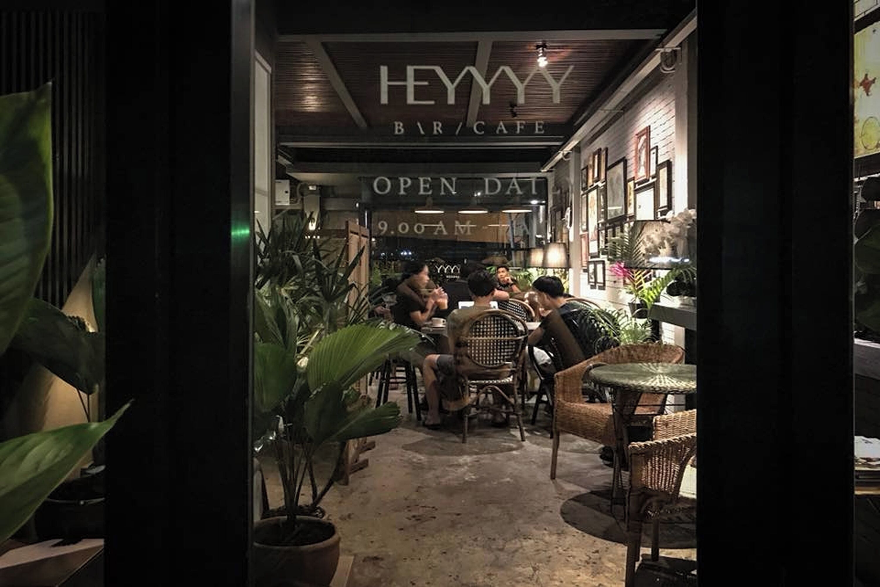 An indoor seating area with wicker furniture and potted plants, under a sign that reads "HEYYY BAR/CAFE" with "OPEN DAILY 9:00 AM" visible.