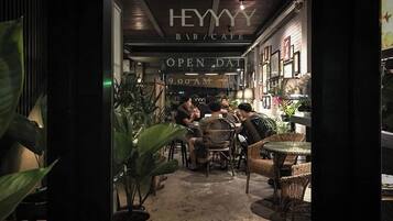 An indoor seating area with wicker furniture and potted plants, under a sign that reads "HEYYY BAR/CAFE" with "OPEN DAILY 9:00 AM" visible.