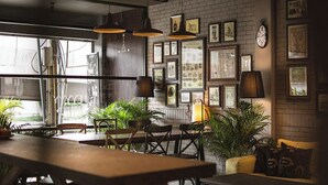 A cozy cafe with wooden tables and chairs, potted plants, and framed pictures on the walls. - Heyyyy Bangkok - Hostel (Bangkok)