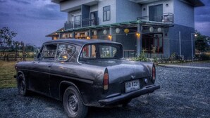 Carro preto antigo estacionado em frente a uma casa moderna de dois andares com varanda e iluminação externa.