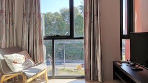 A room with a large window, a sofa with a patterned cushion, a TV on a stand, and a view of greenery outside. - Nice Nap Hostel (Sa Khu)