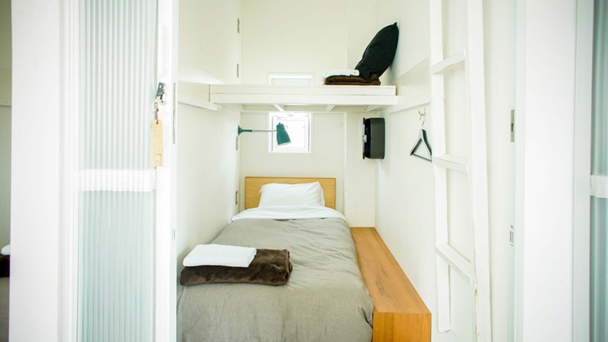 A compact room with a bed, a small desk, a shelf with a black object, and a window with blinds.