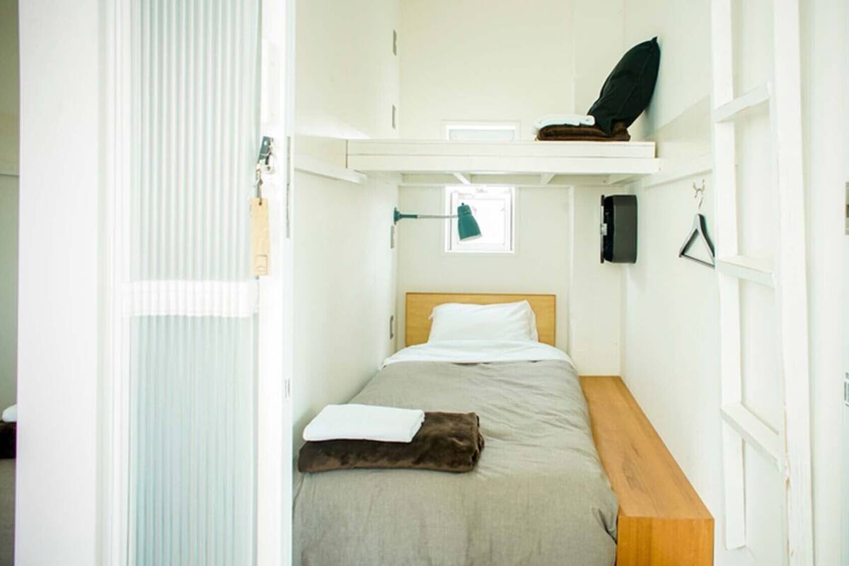 A compact room with a bed, a small desk, a shelf with a black object, and a window with blinds.