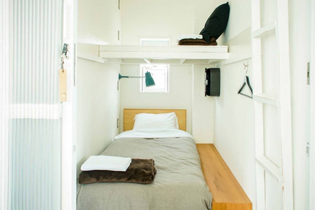 A small, narrow room with a single bed, a wooden bench, a small table, and a window.