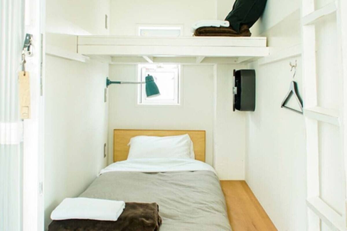 A small, single-bed room with a wooden headboard, a window, a shelf with a black bag, and a hanging coat hook.