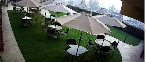 An outdoor seating area with multiple tables and chairs under large umbrellas, arranged on artificial grass.