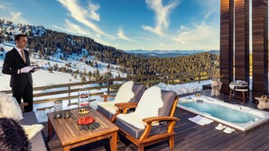 Een houten terras met een jacuzzi, twee stoelen, een tafel met lekkernijen en uitzicht op een besneeuwd berglandschap.