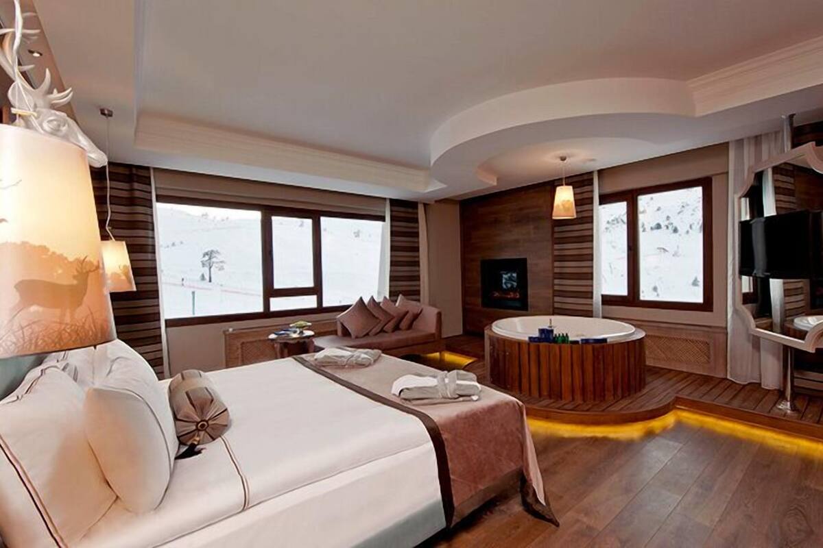 Una habitación de hotel moderna con una cama grande, una zona de estar con un sofá y una mesita, una bañera redonda de madera y ventanas amplias con vistas nevadas.
