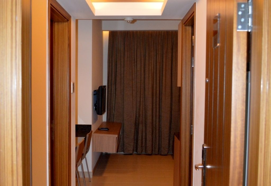 Un pasillo de hotel con puertas de madera, una lámpara en el techo, un televisor de pantalla plana montado en la pared y una cortina al final.
