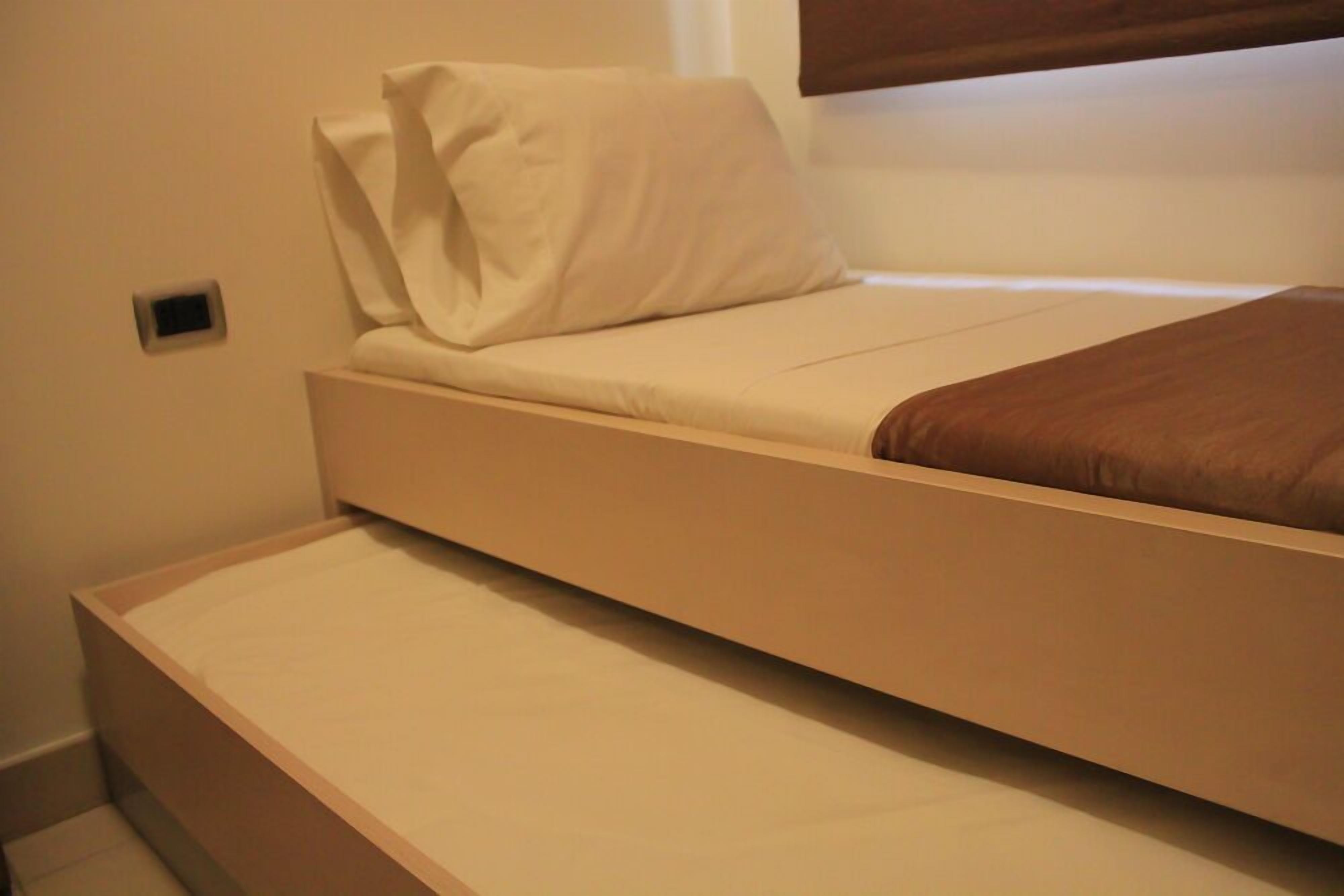 A single bed with a mattress and a pillow, a small bedside table, and a window with blinds.