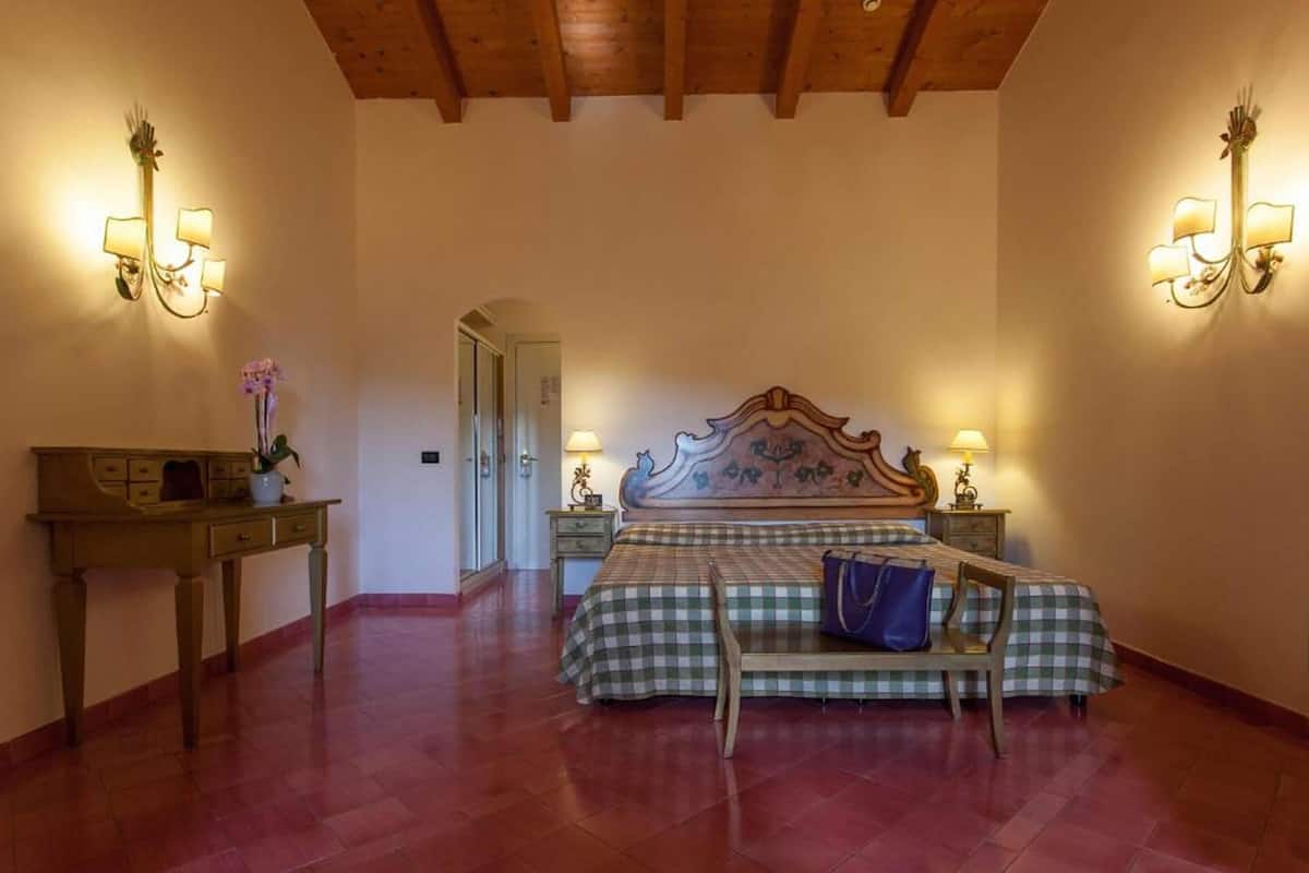 A bedroom with a wooden ceiling, a checkered bedspread, two bedside lamps, a nightstand, and a desk with a vase of flowers.
