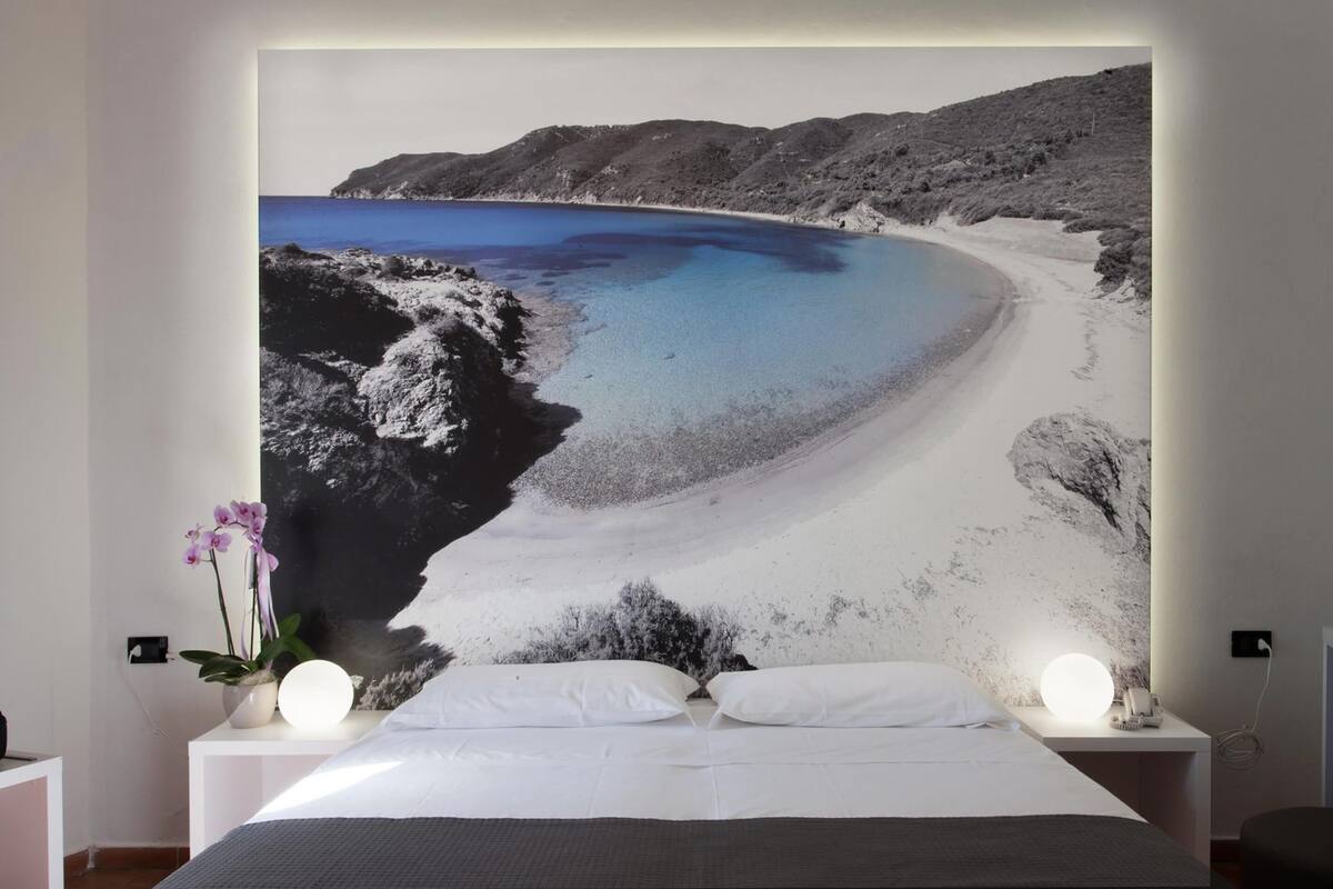 A bedroom with a large bed, white bedding, a nightstand with a lamp, and a wall-mounted TV displaying a beach scene.