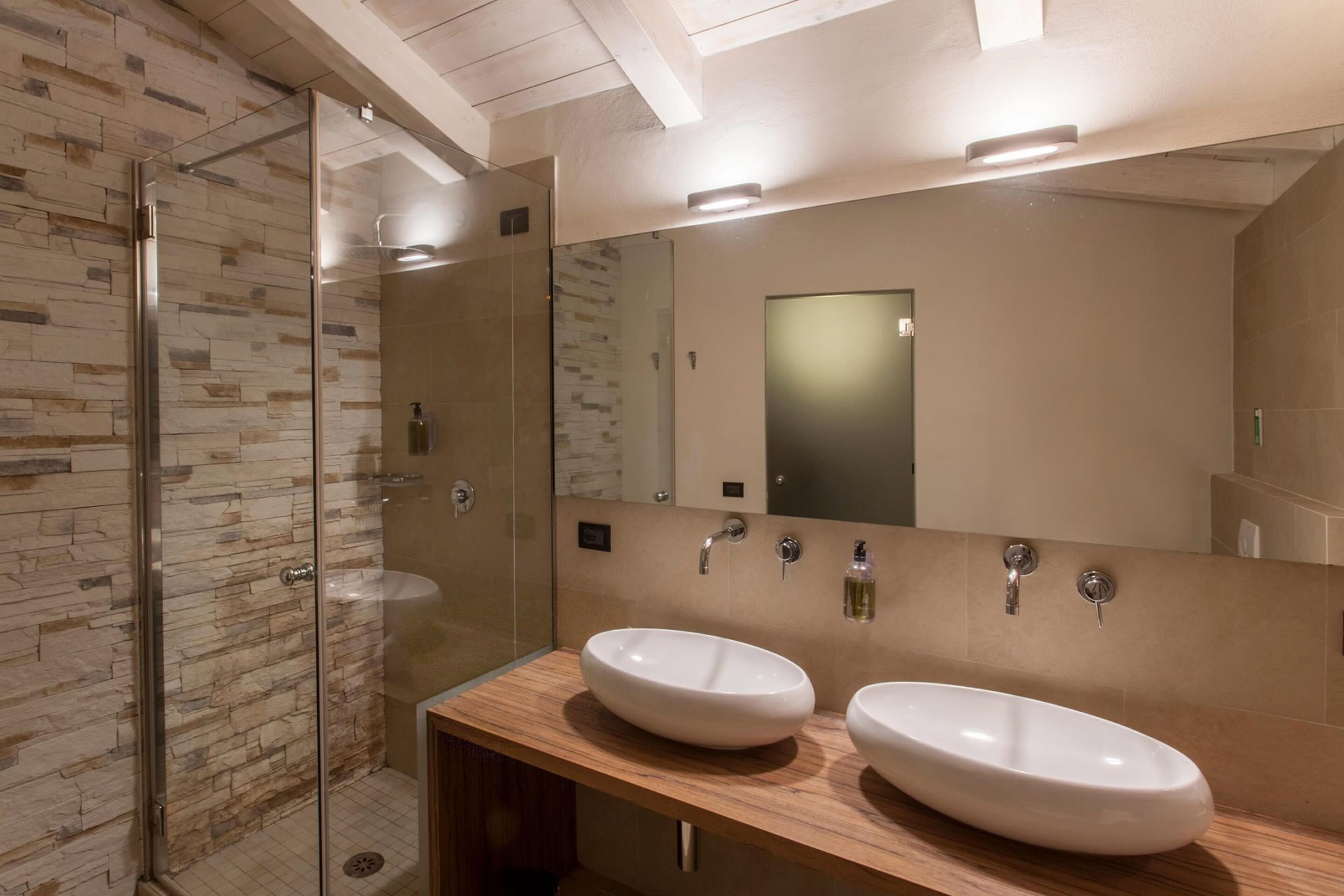 Un baño moderno con cabina de ducha de vidrio, dos lavamanos blancos y un espejo grande sobre los lavamanos.