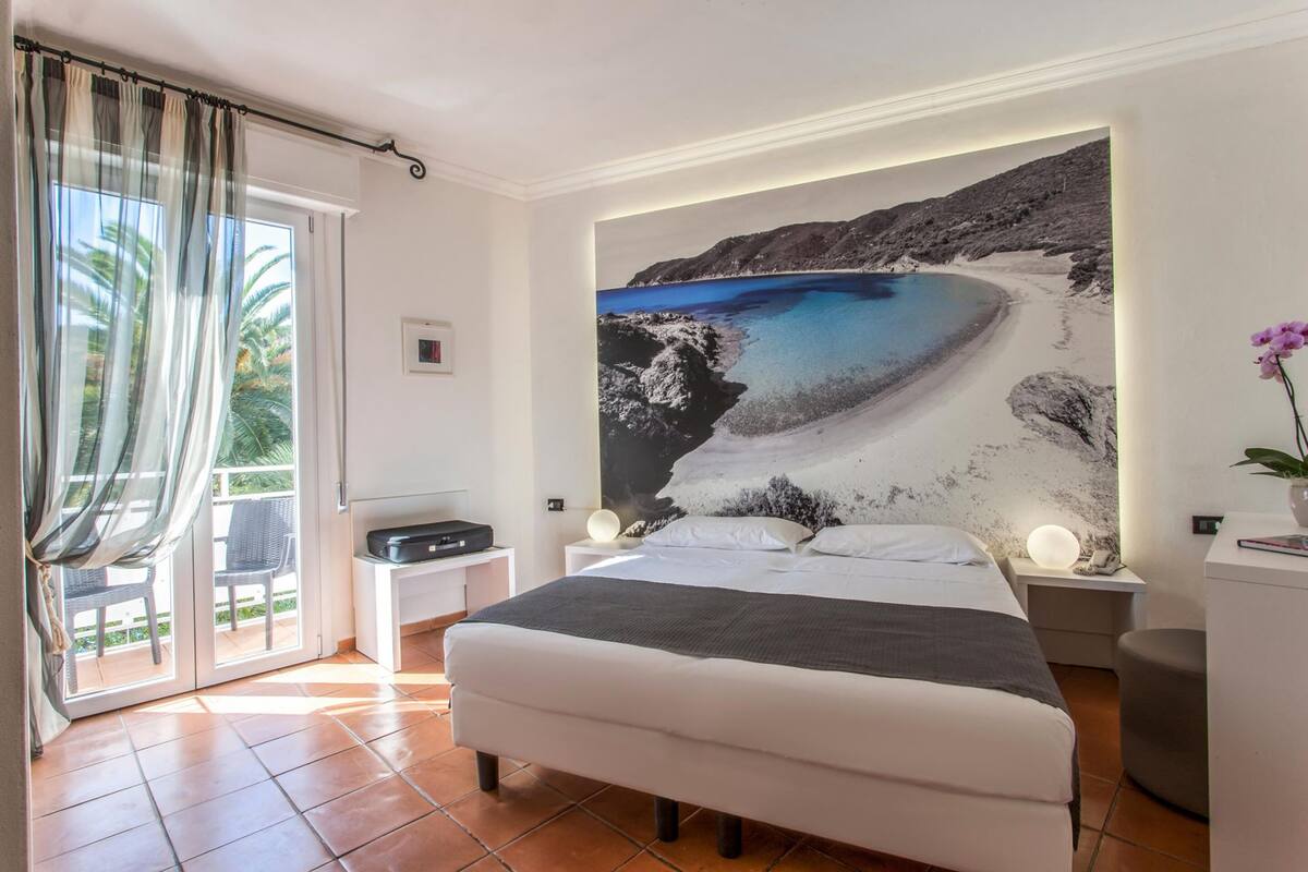 A bedroom with a large bed, a view of a beach through a window, a small table, and a nightstand with a lamp.