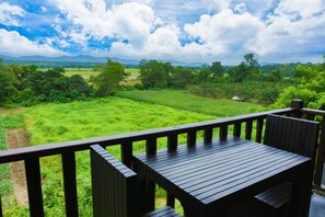 Una terraza de madera con una mesa y sillas que da a un exuberante paisaje verde con árboles y montañas distantes bajo un cielo parcialmente nublado.