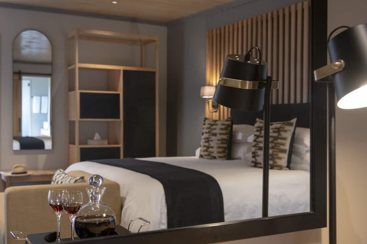 A hotel room with a neatly made bed, a bedside table with glasses and a decanter, a bookshelf, and a large mirror.