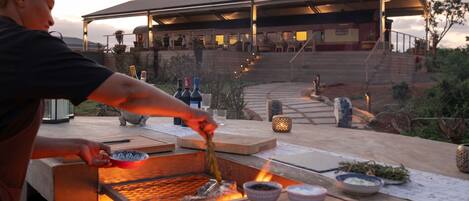 A chef grilling food outdoors with a view of a building in the background.