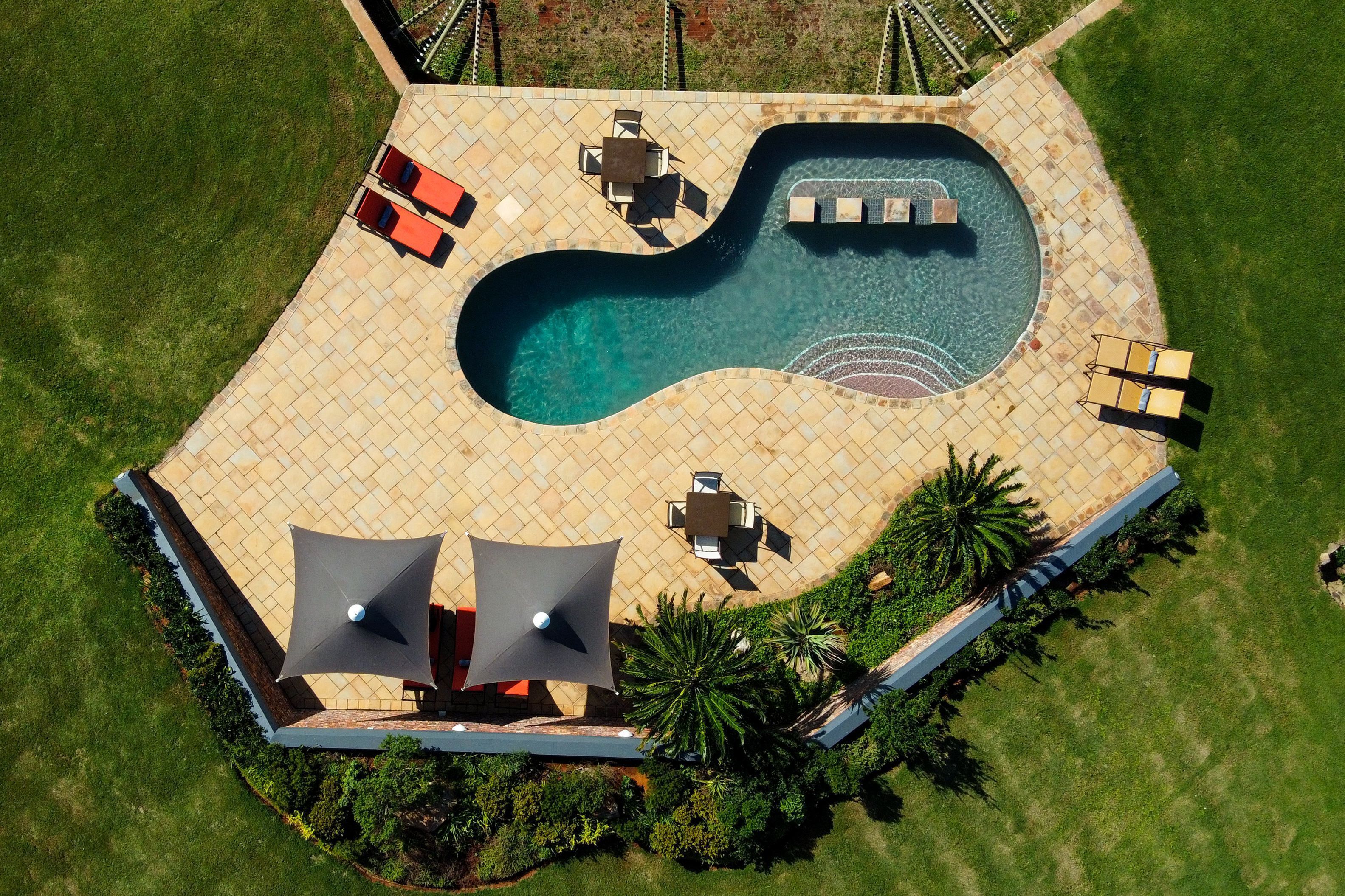 Vue aérienne d’un espace piscine à la forme originale, entouré d’une terrasse carrelée, de coins salon extérieurs et de palmiers.
