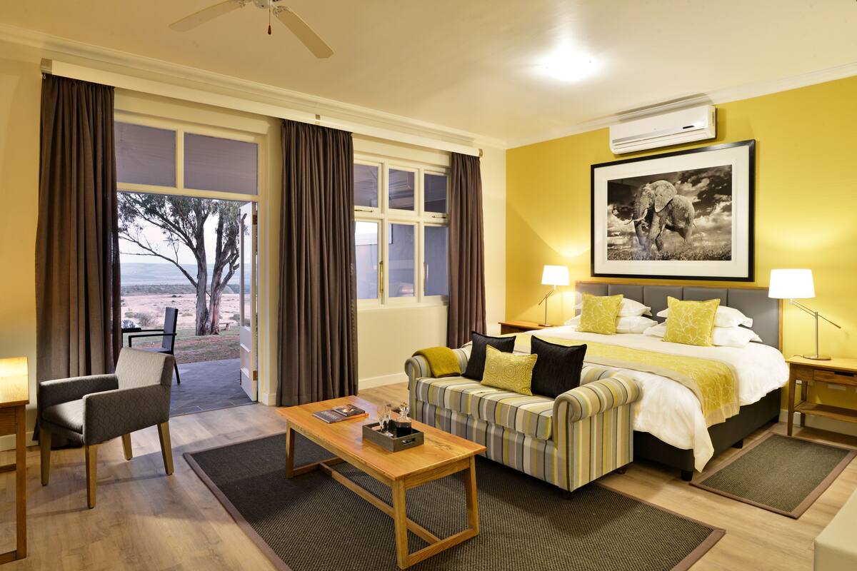 A hotel room with a bed, a sofa, a coffee table, and a chair. There is a window with curtains, a picture on the wall, and an air conditioning unit.