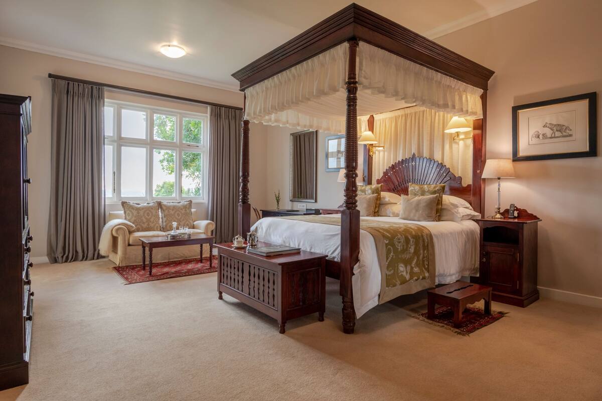 A four-poster bed with a canopy, a sitting area with a sofa and armchair, a bedside table with a lamp, and a framed picture on the wall.